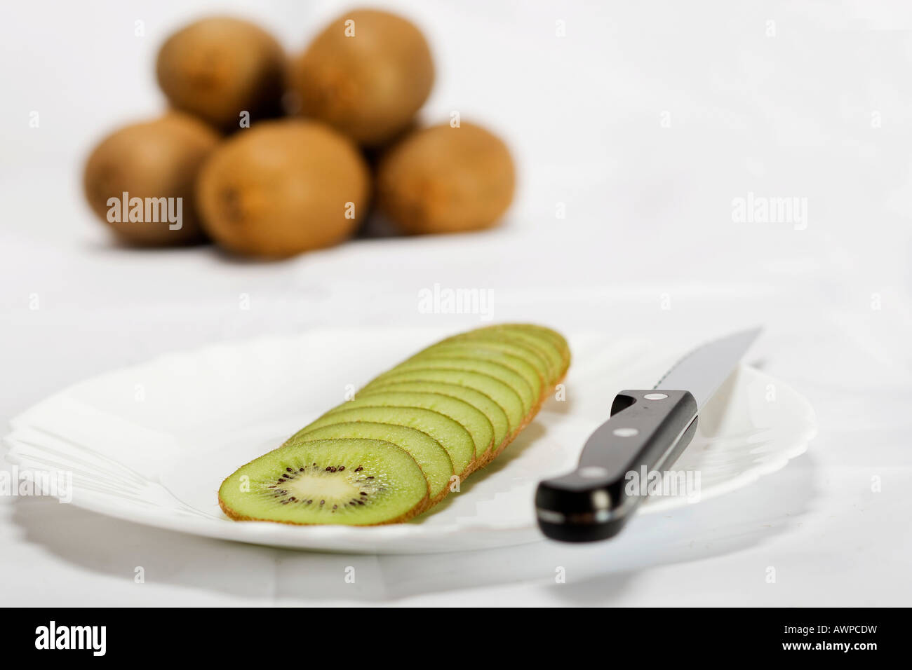 Single sliced kiwi on a plate with knife, more kiwis piled at the back Stock Photo