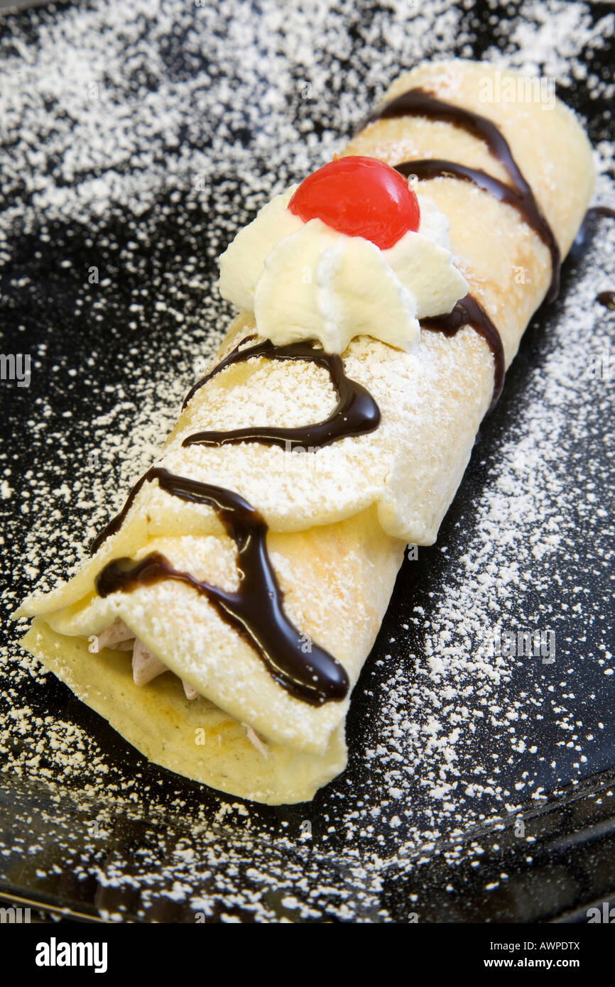 Crepe filled with cream and topped with icing sugar, chocolate glaze, whipped cream and a maraschino cherry Stock Photo