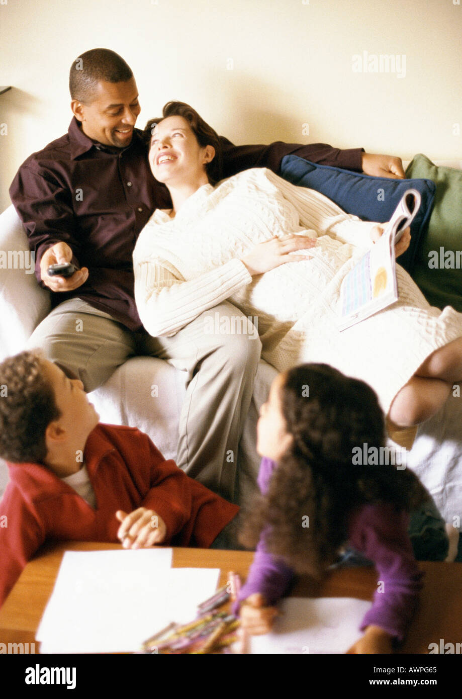 Man and pregnant woman on sofa, children looking over shoulders at them Stock Photo