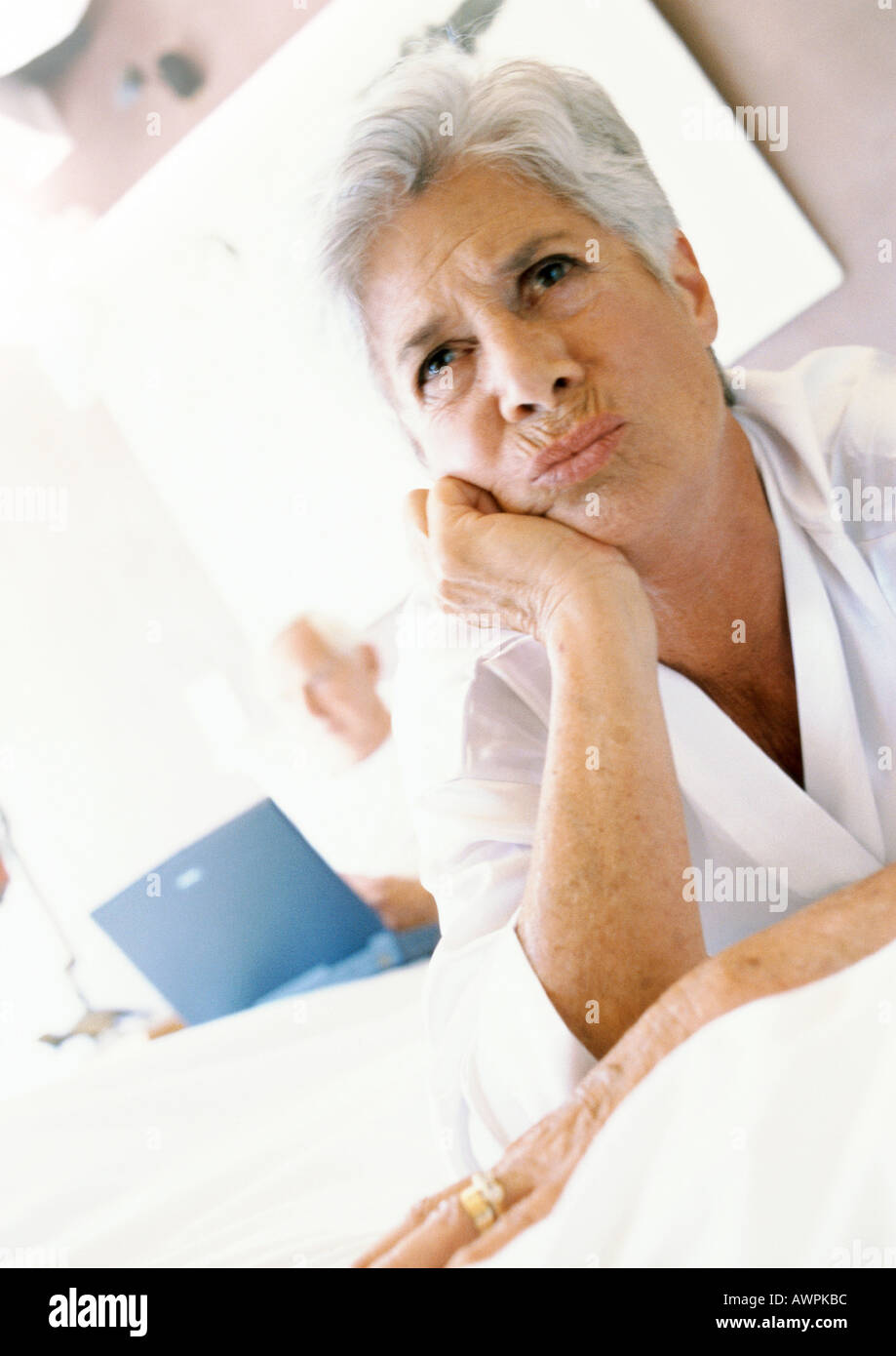 Senior woman with hand under chin, frowning, man using laptop in background Stock Photo