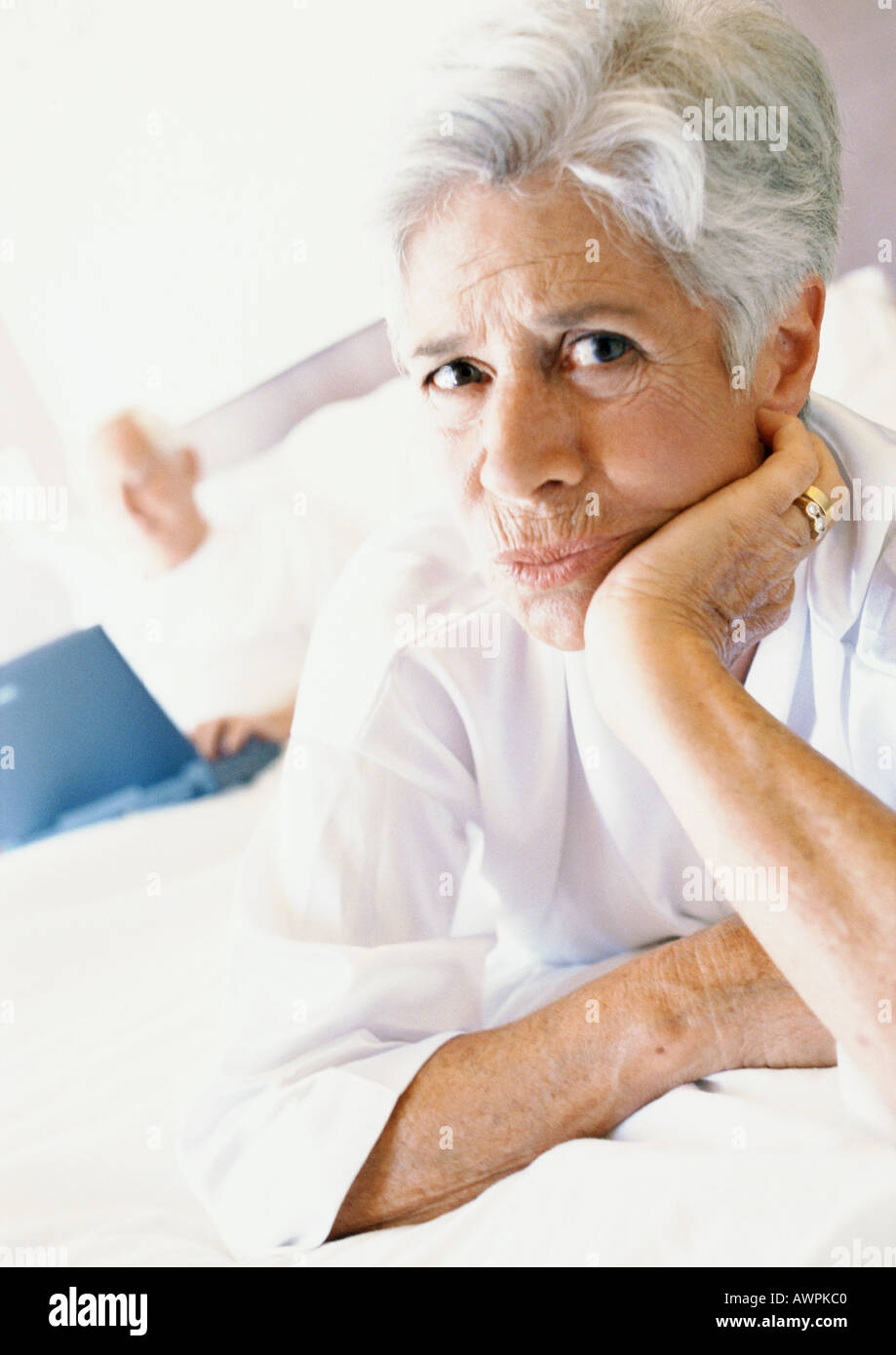 Senior woman on bed with hand under chin, man using laptop in background Stock Photo