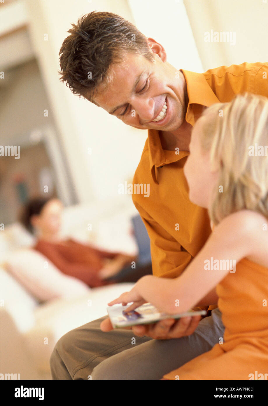 Father and daughter looking at book, smiling, woman sitting on sofa in background Stock Photo