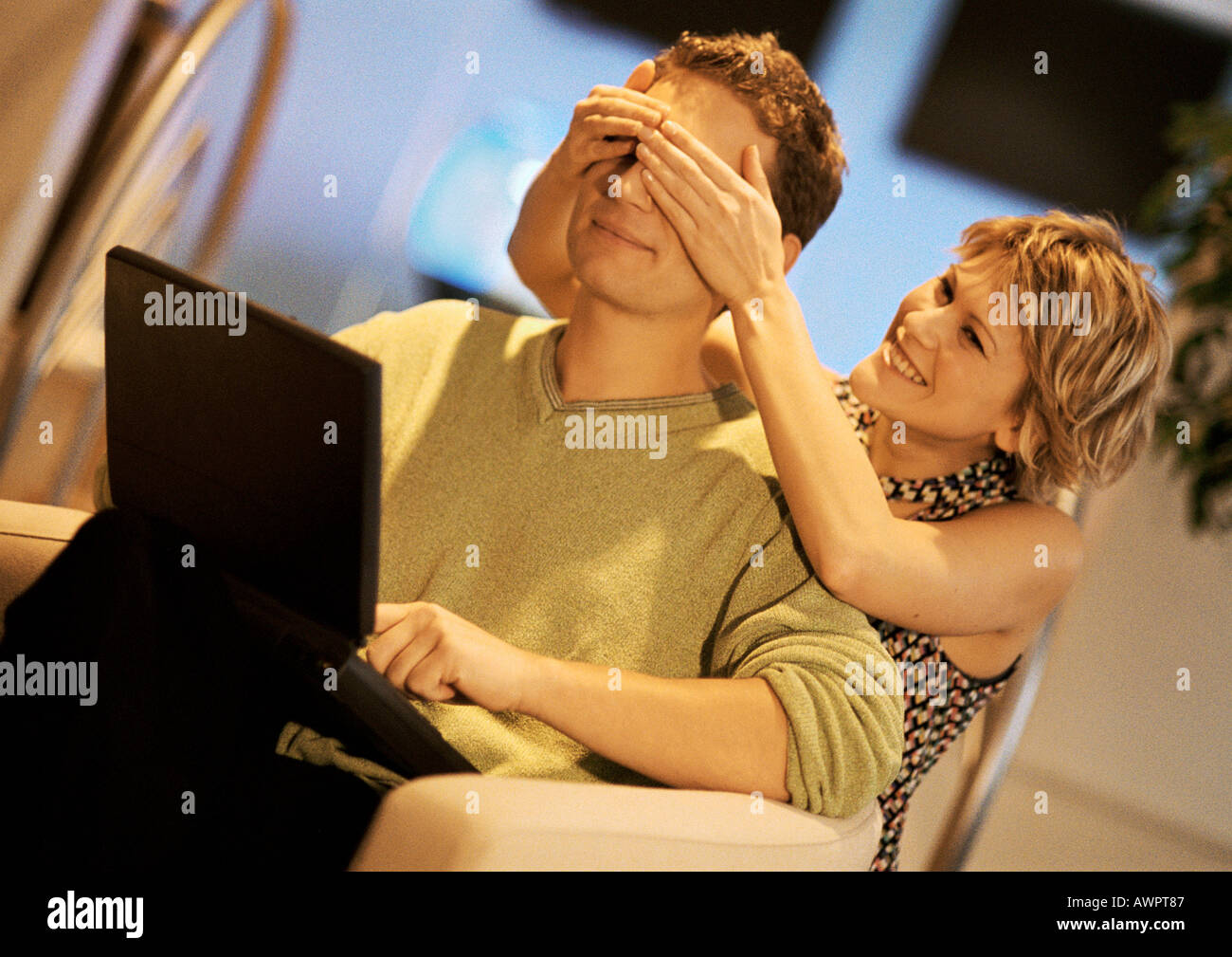 Couple, man using laptop, woman covering man's eyes from behind Stock Photo
