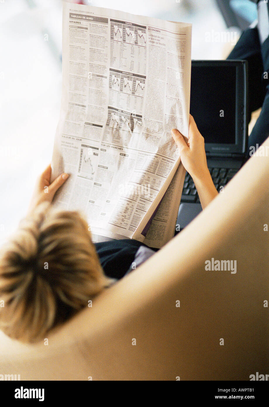 Woman reading newspaper, laptop on lap, rear view, elevated angle. Stock Photo