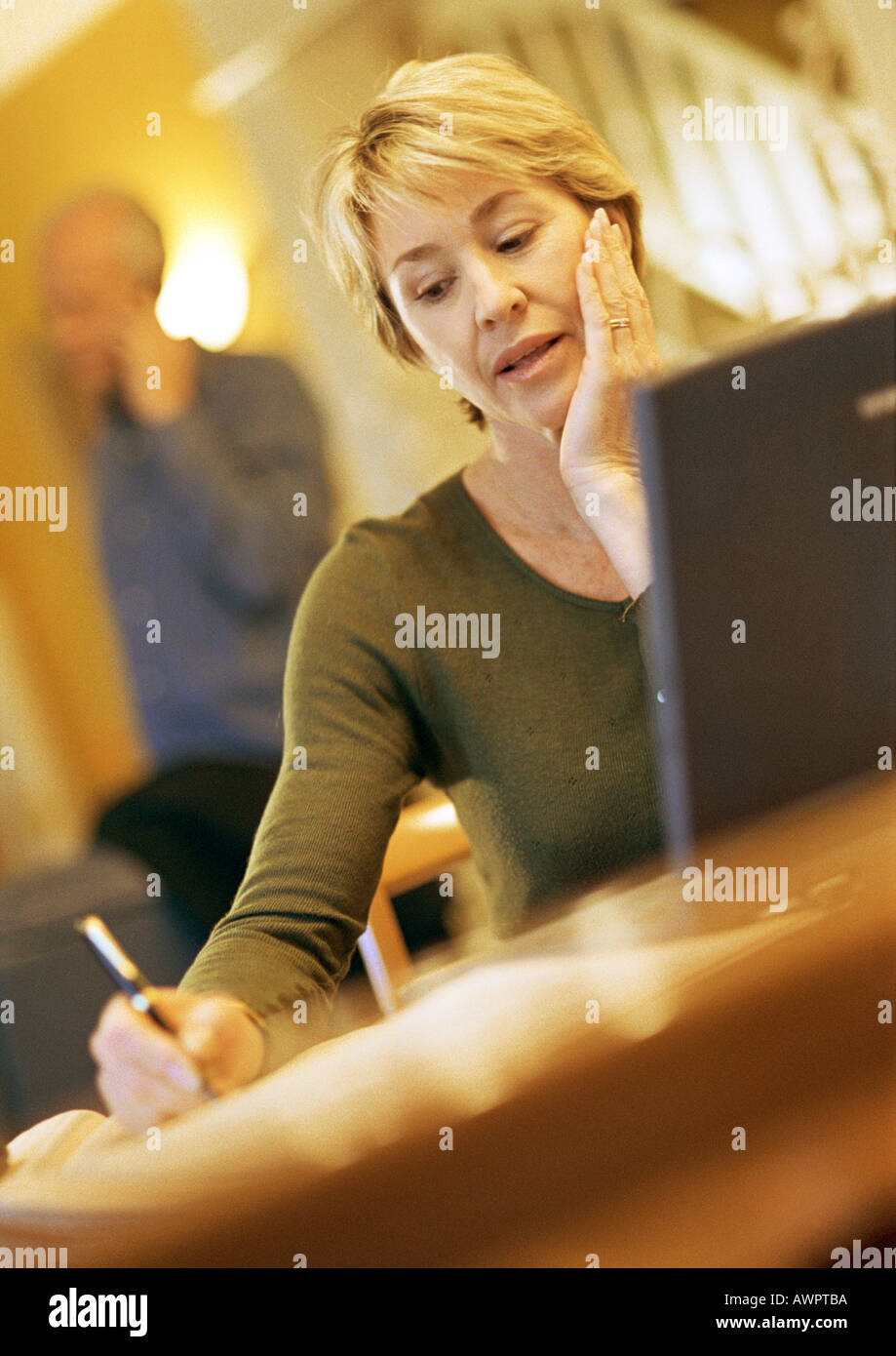 Mature woman using laptop, taking notes, man using cell phone in background Stock Photo