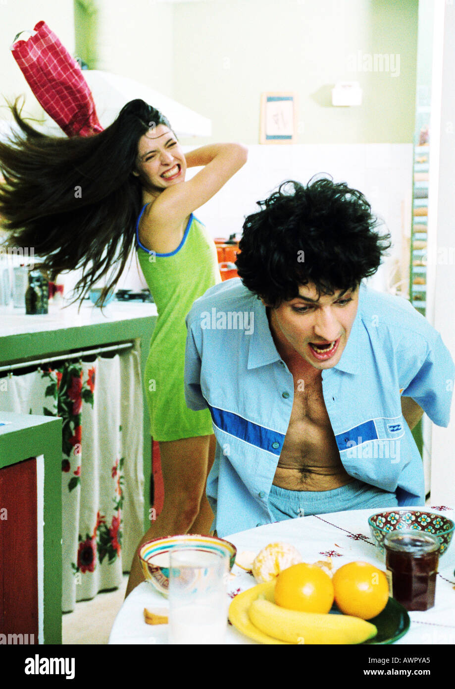 Man at breakfast table, woman swinging kitchen towel at man. Stock Photo