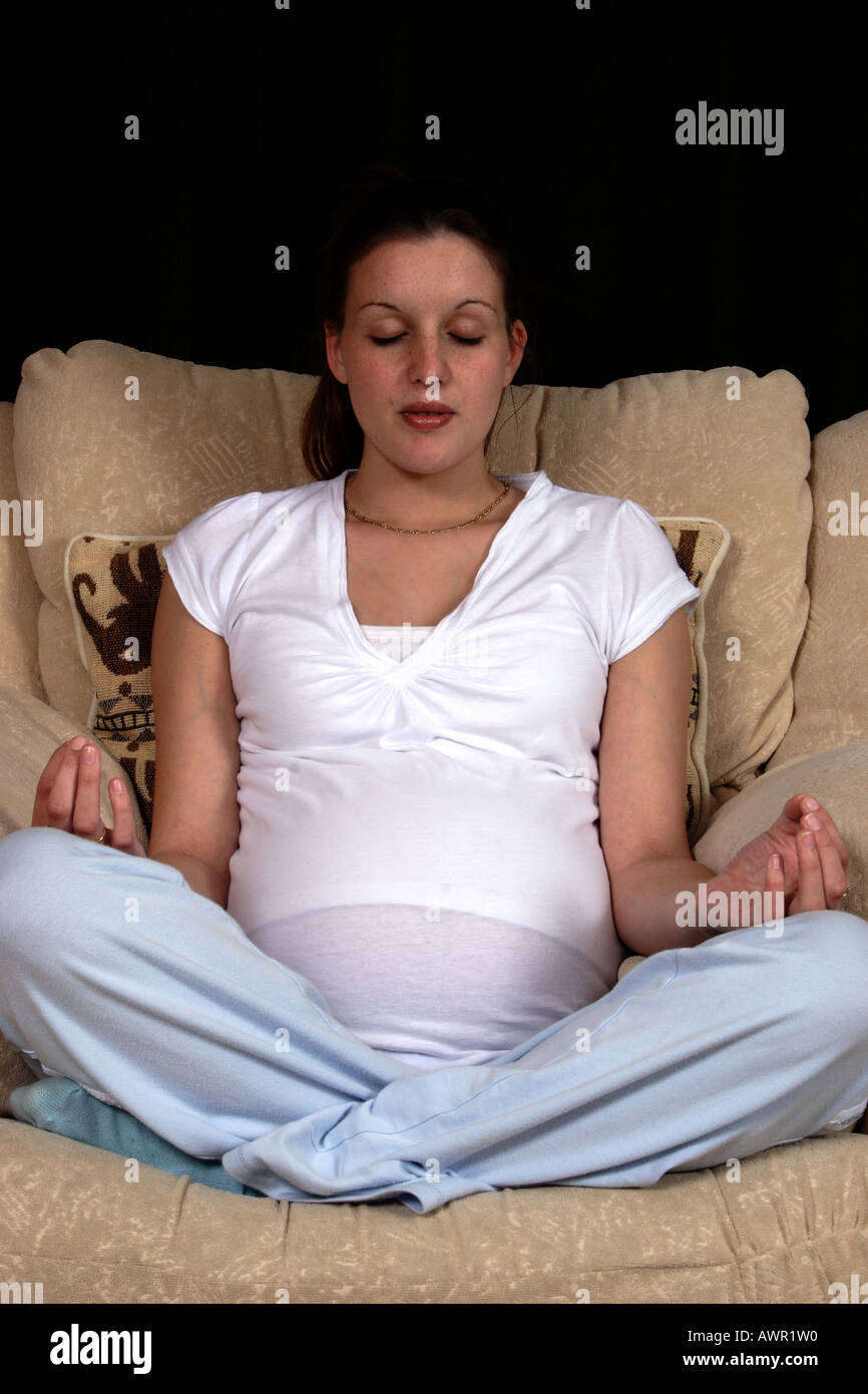 Young heavily pregnant woman sitting on armchair in yoga position meditating Stock Photo