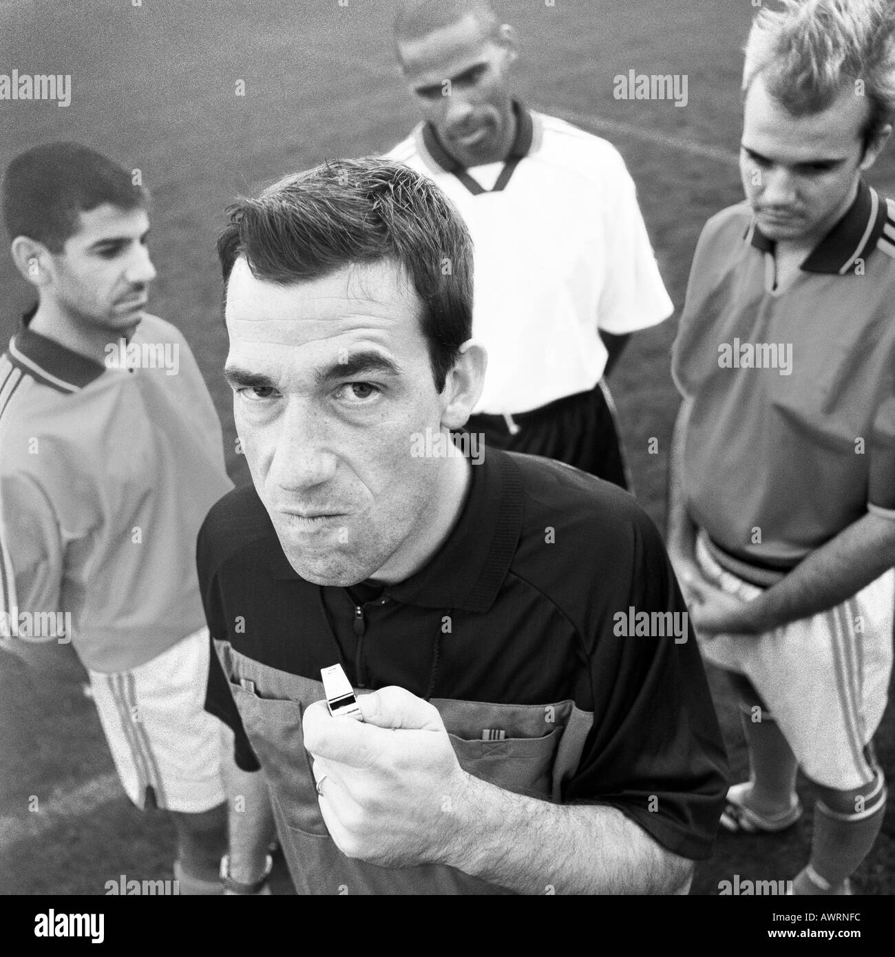 Referee holding whistle in front of three players, looking into camera, b&w. Stock Photo