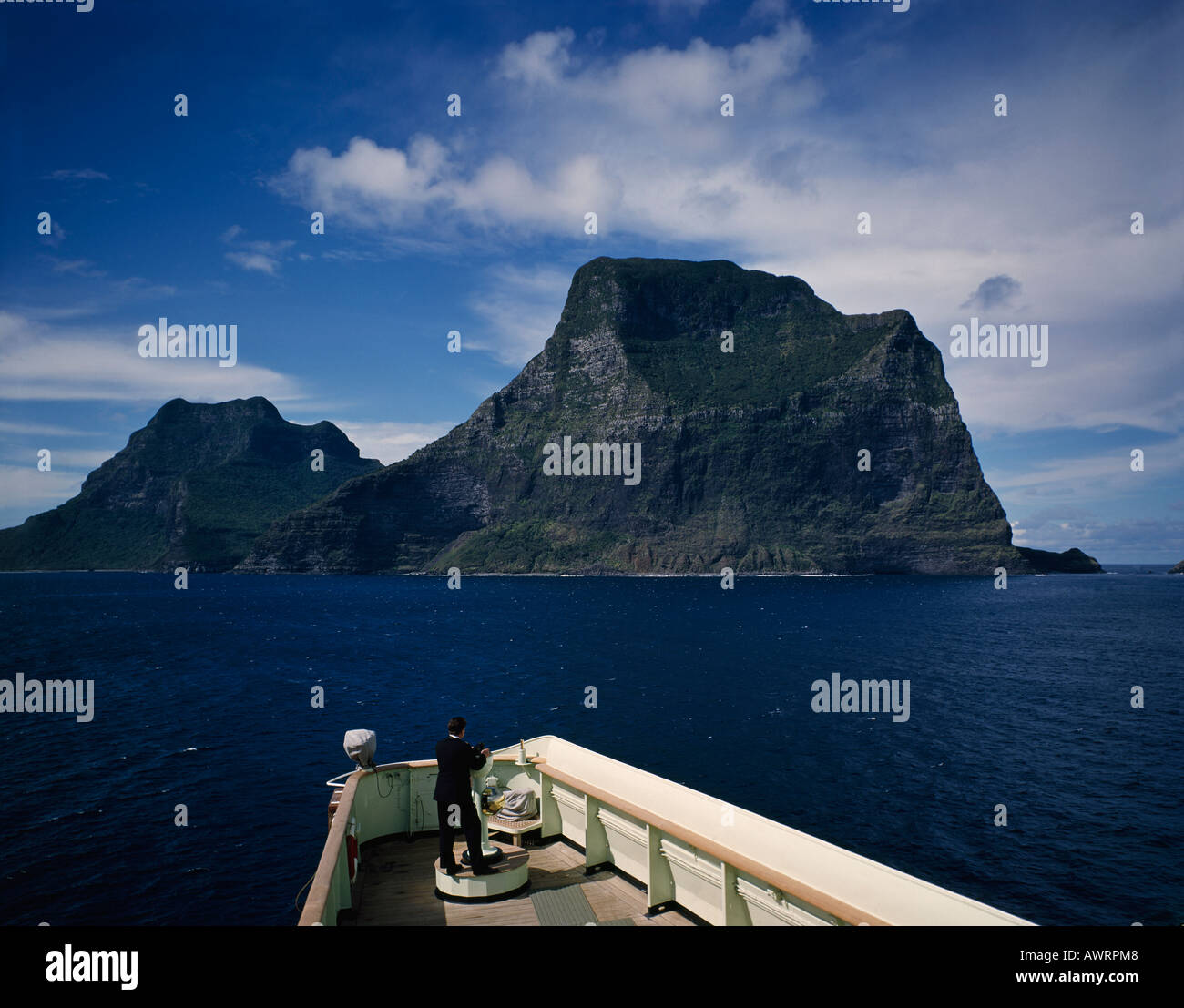 Lone officer on bridge of ship looking across ocean to majestic cliffs of green forested Lord Howe Island in the South Pacific Stock Photo