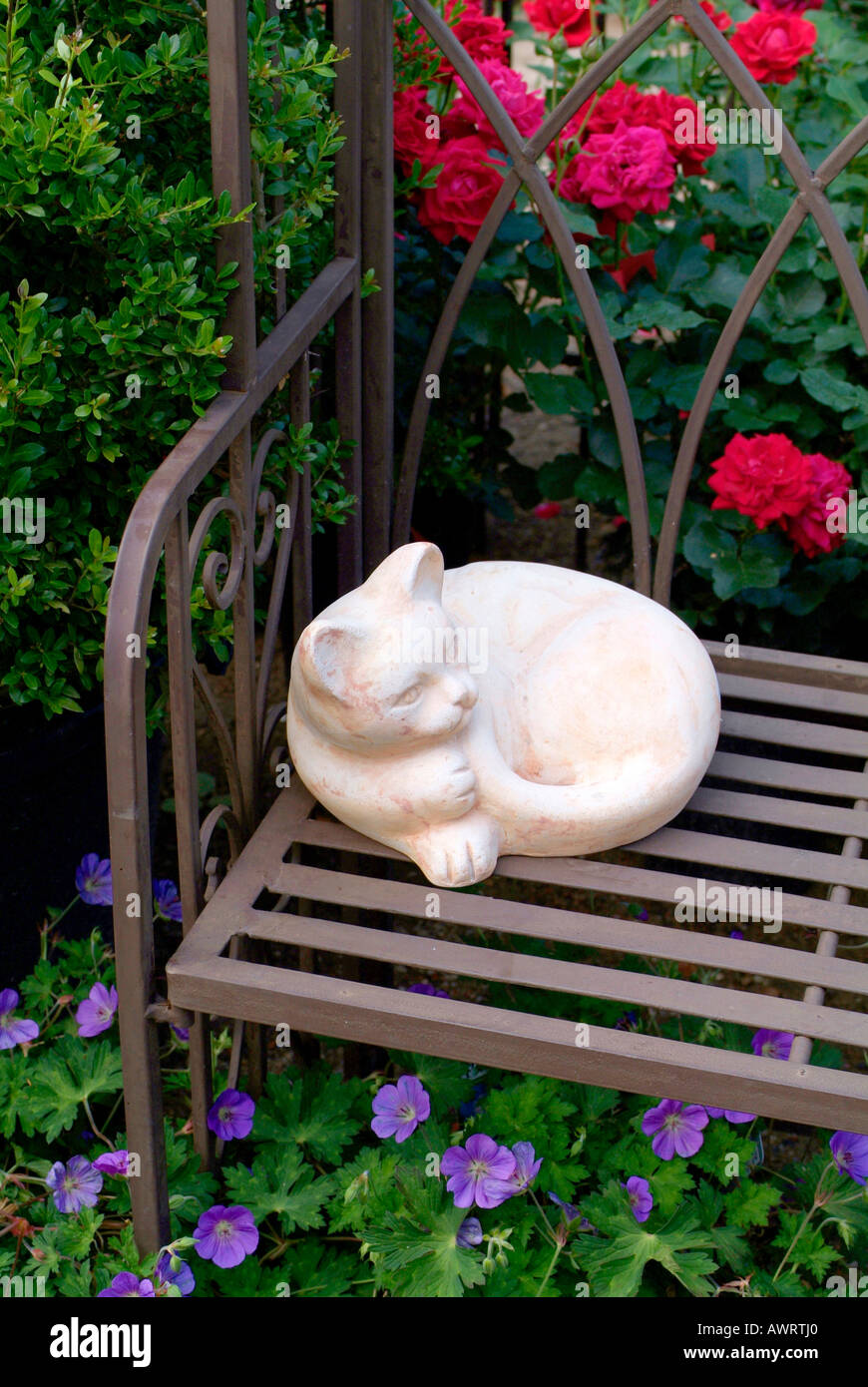 Garden deco, stone cat is lying on a metal bench in front of buxus, roses rosa and geranium Stock Photo