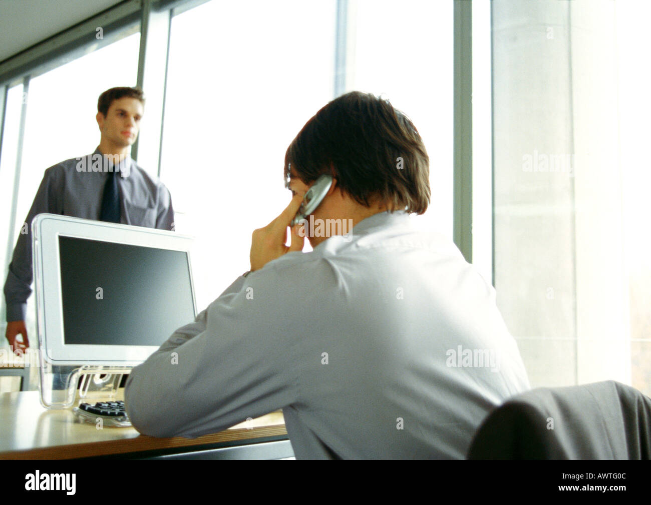 Businessman sitting at computer, using cell phone, second businessman walking in office Stock Photo