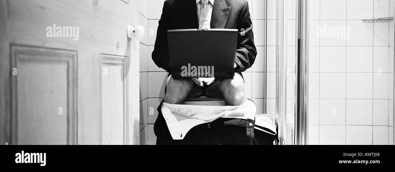 Businessman sitting on toilet with pants down, using laptop, mid-section, b&w, panoramic view Stock Photo