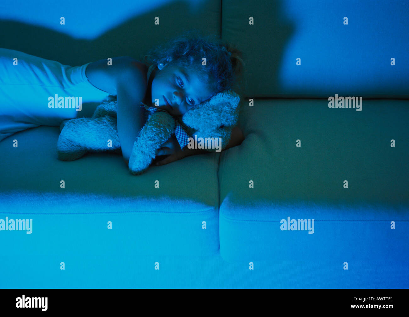 Little girl lying on sofa cuddling with teddy bear, blue light of television Stock Photo