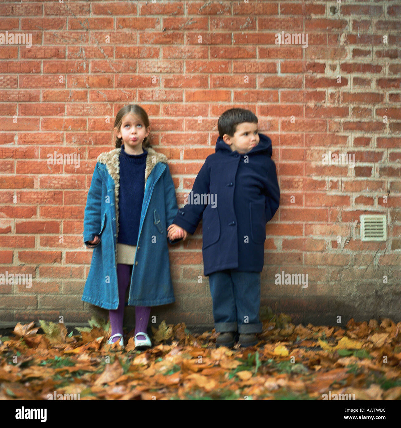 Boy and girl holding hands and making faces in front of brick wall, full length Stock Photo