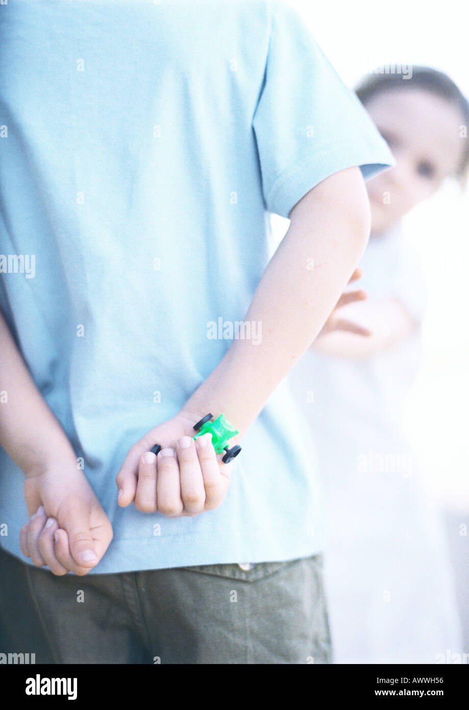 Child with hands behind back, one holding toy car, close-up, girl in background reaching Stock Photo