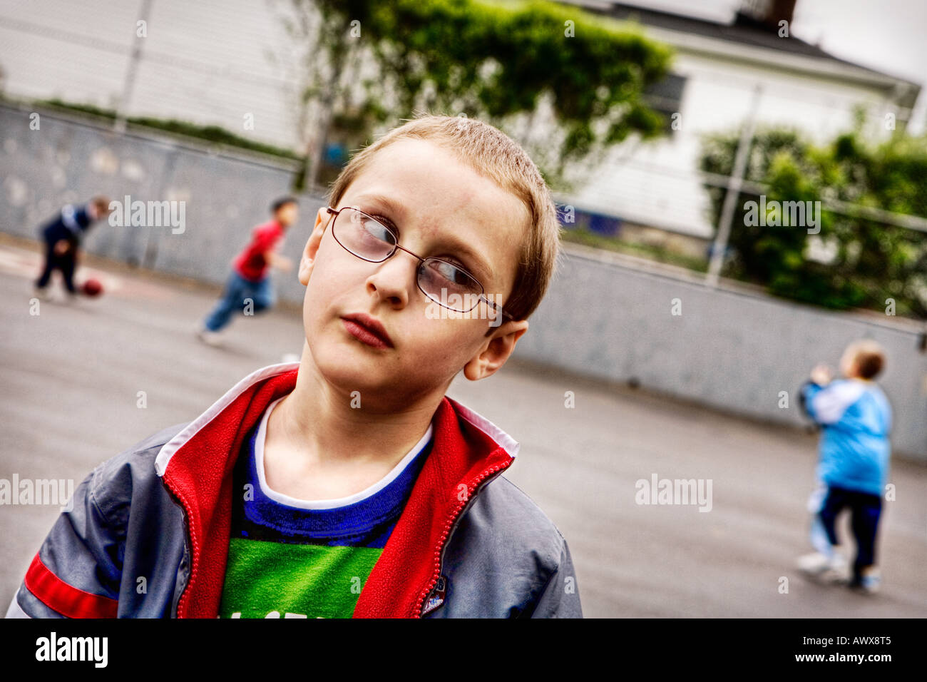 young boy wearing glasses standing on a playground rolling his eyes while other kids are playing ball behind in the background Stock Photo