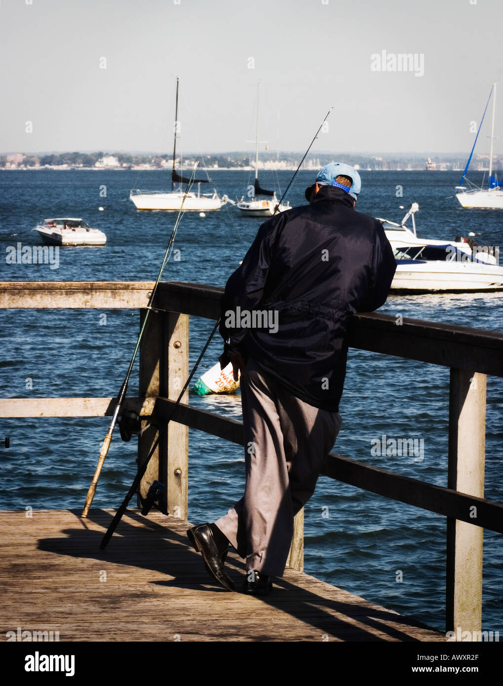 man fishing off a wooden pier with water in background and many boats anchored at a marina Stock Photo