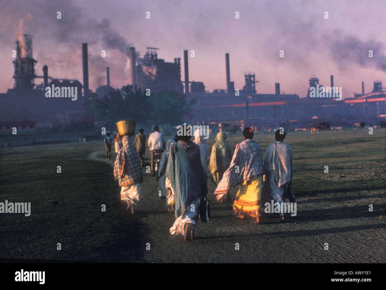 India women in saris going to work at industrial power plant polluting the sky with smoke at sunrise Stock Photo