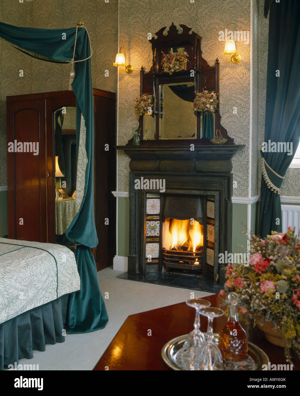 Carved Victorian overmantel mirror above fireplace with patterned tiles in bedroom with Arts Crafts wallpaper Stock Photo