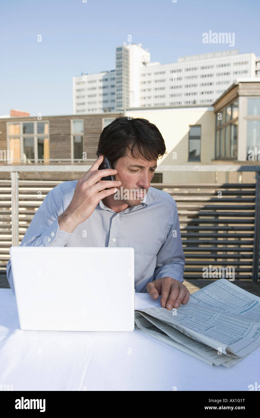 A businessman sitting on a rooftop terrace with a newspaper, mobile phone and laptop Stock Photo