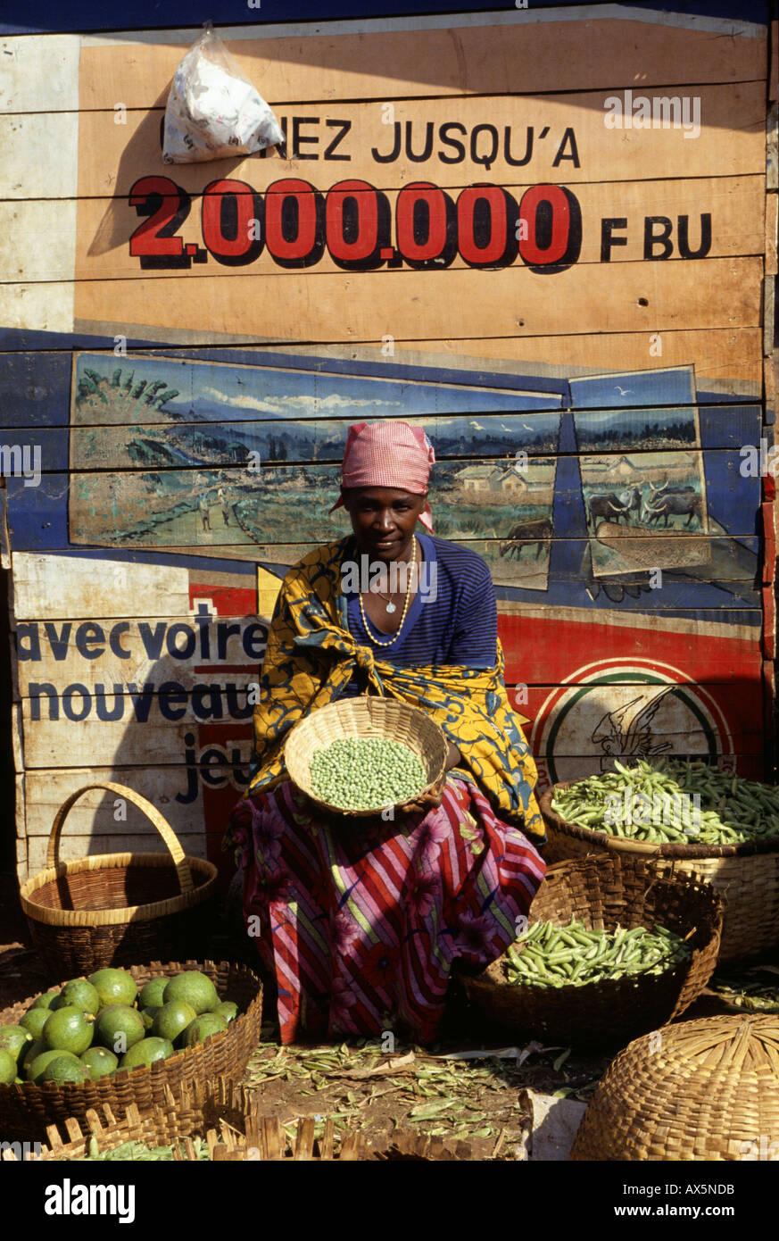 Gitega, Burundi. Woman in the market selling peas and fruit in front of a painted wall with pictures; French words. Stock Photo