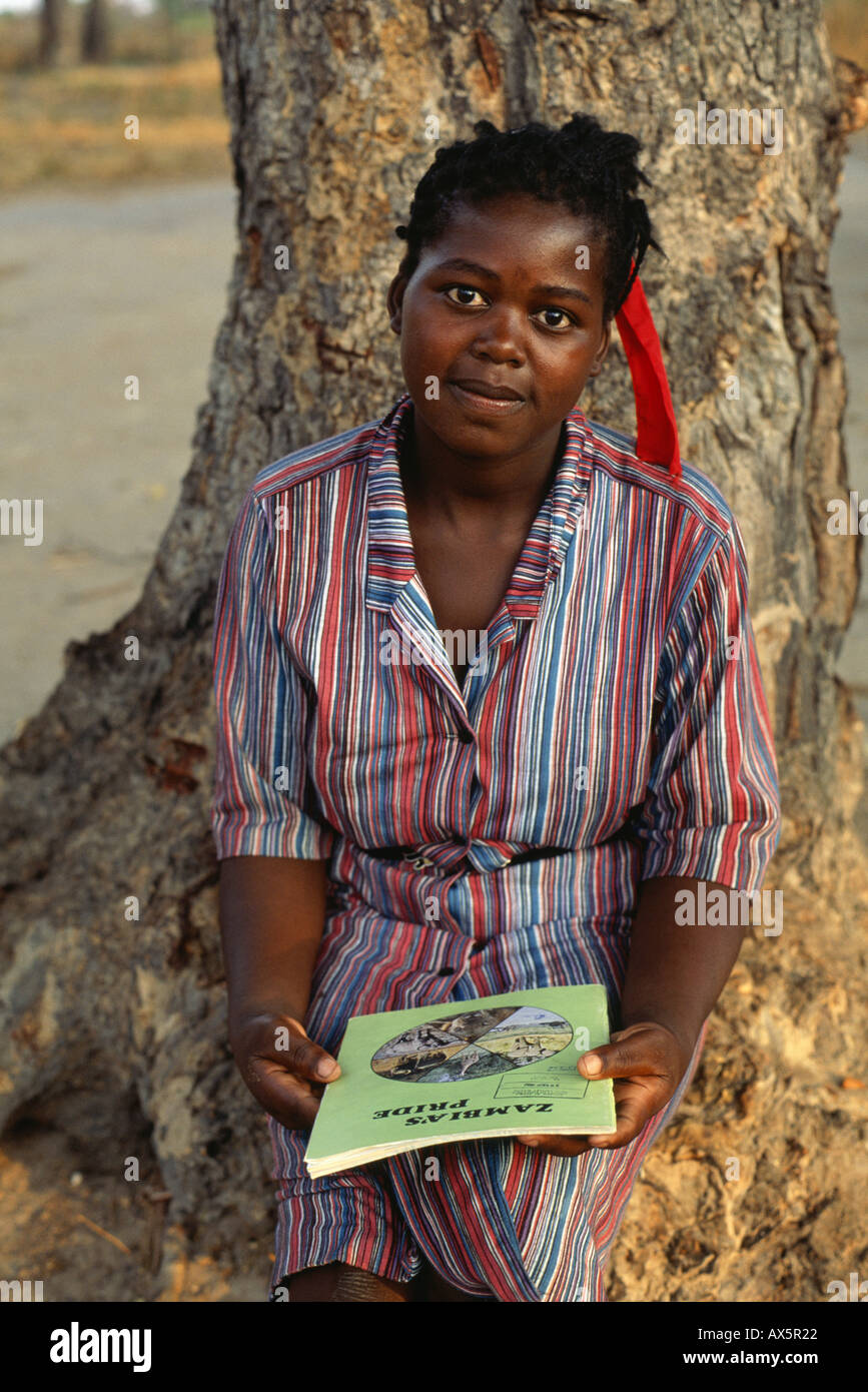 Mbati, Zambia. Woman student in uniform holding a booklet called "Zambia's Pride", sitting at the foot of a tree. Stock Photo