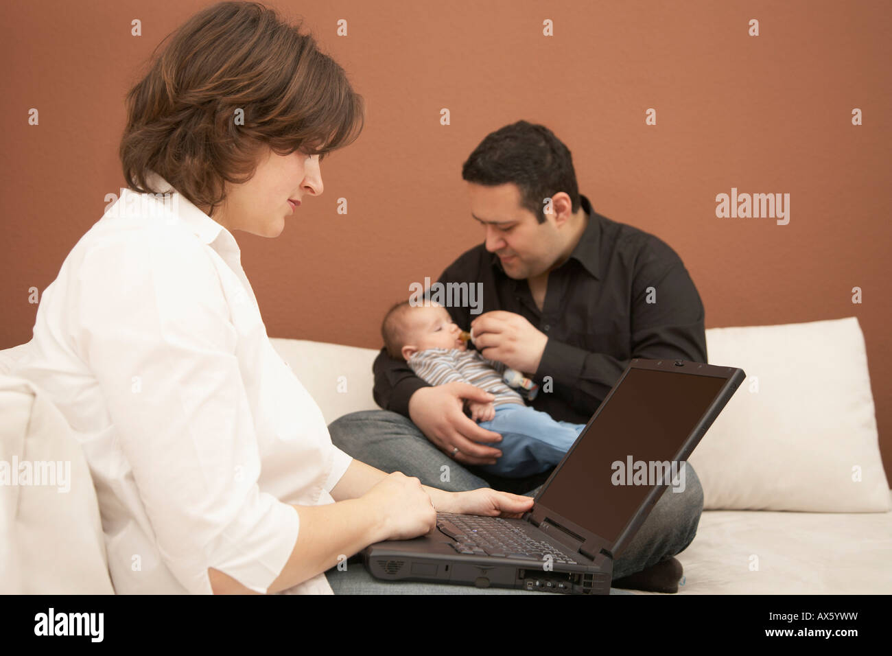 Young woman working on laptop while her husband cares for their baby Stock Photo