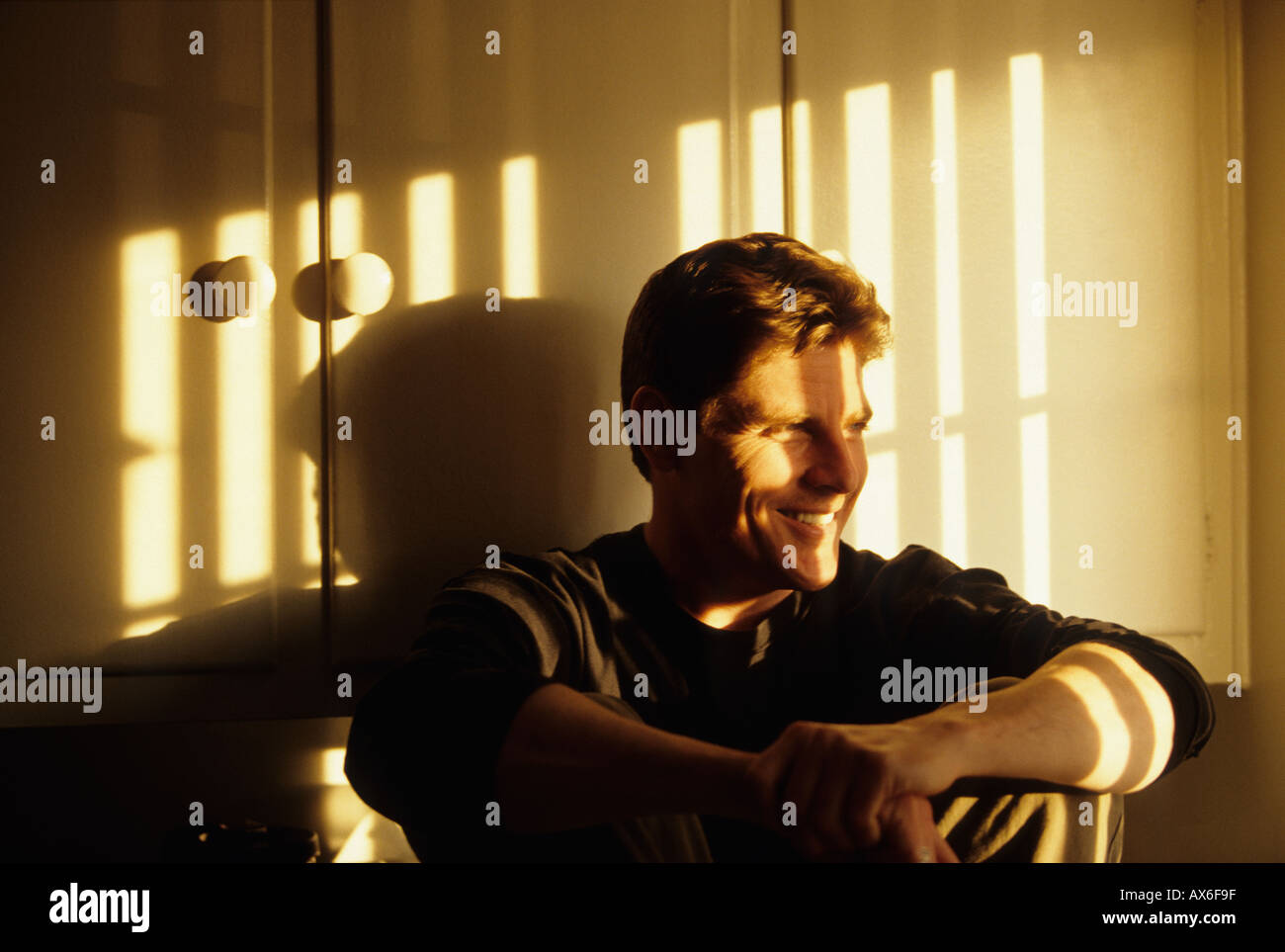 Man sitting on the floor in front of cabinets smiling with shadow from vertical blinds at sunset across his face Stock Photo