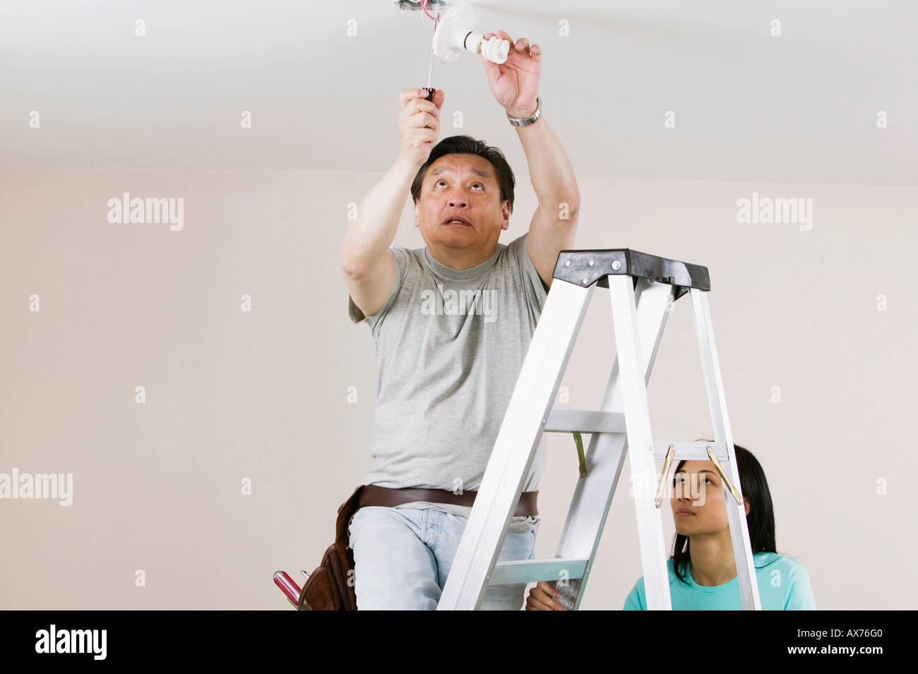 Low angle view of a mid adult man fixing a light fixture with a young woman holding a ladder Stock Photo