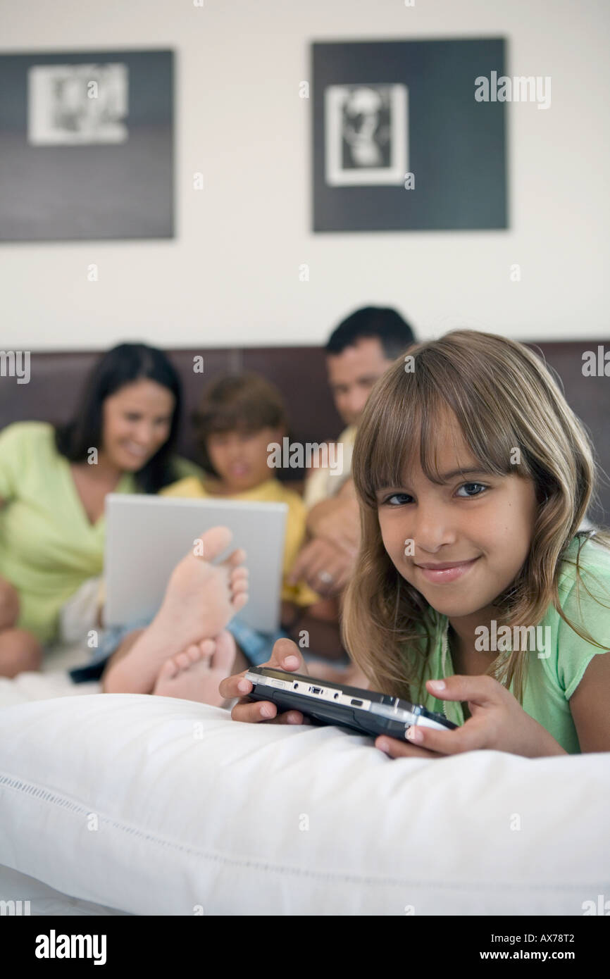 Girl holding a handheld video game with her parents and brother looking at a laptop in the background Stock Photo