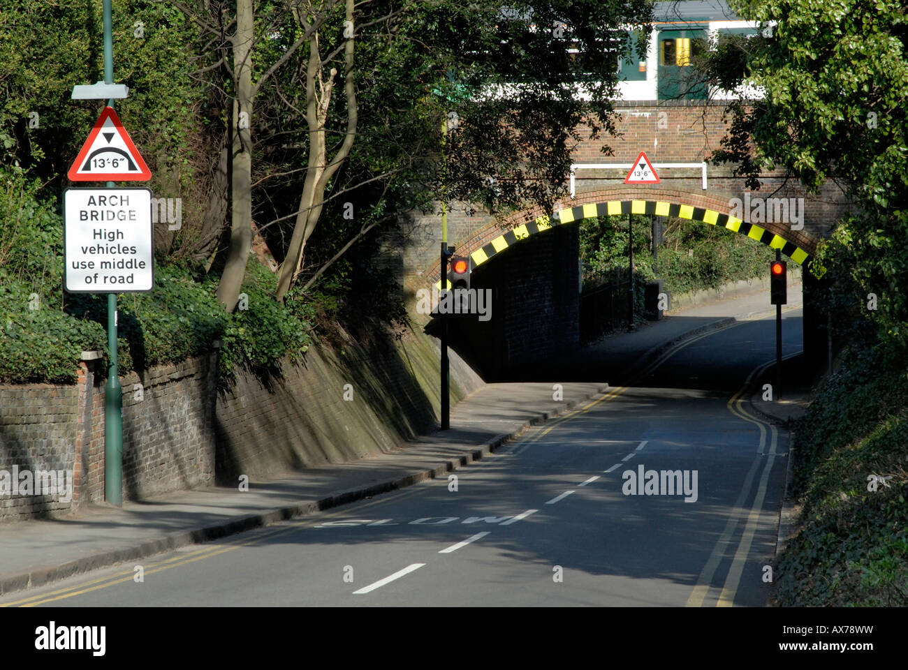 Low arch bridge with warning sign and high visibility markings, with passenger train above, Cheam, south London, Surrey, England Stock Photo