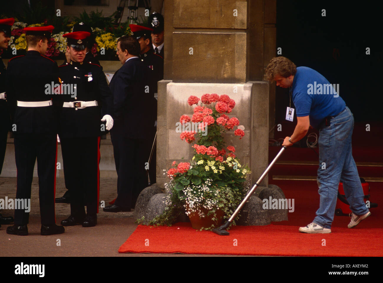 A civilian cleaner hoovers an official red carpet under the watchful eyes of guardsmen at St James' Palace. Stock Photo