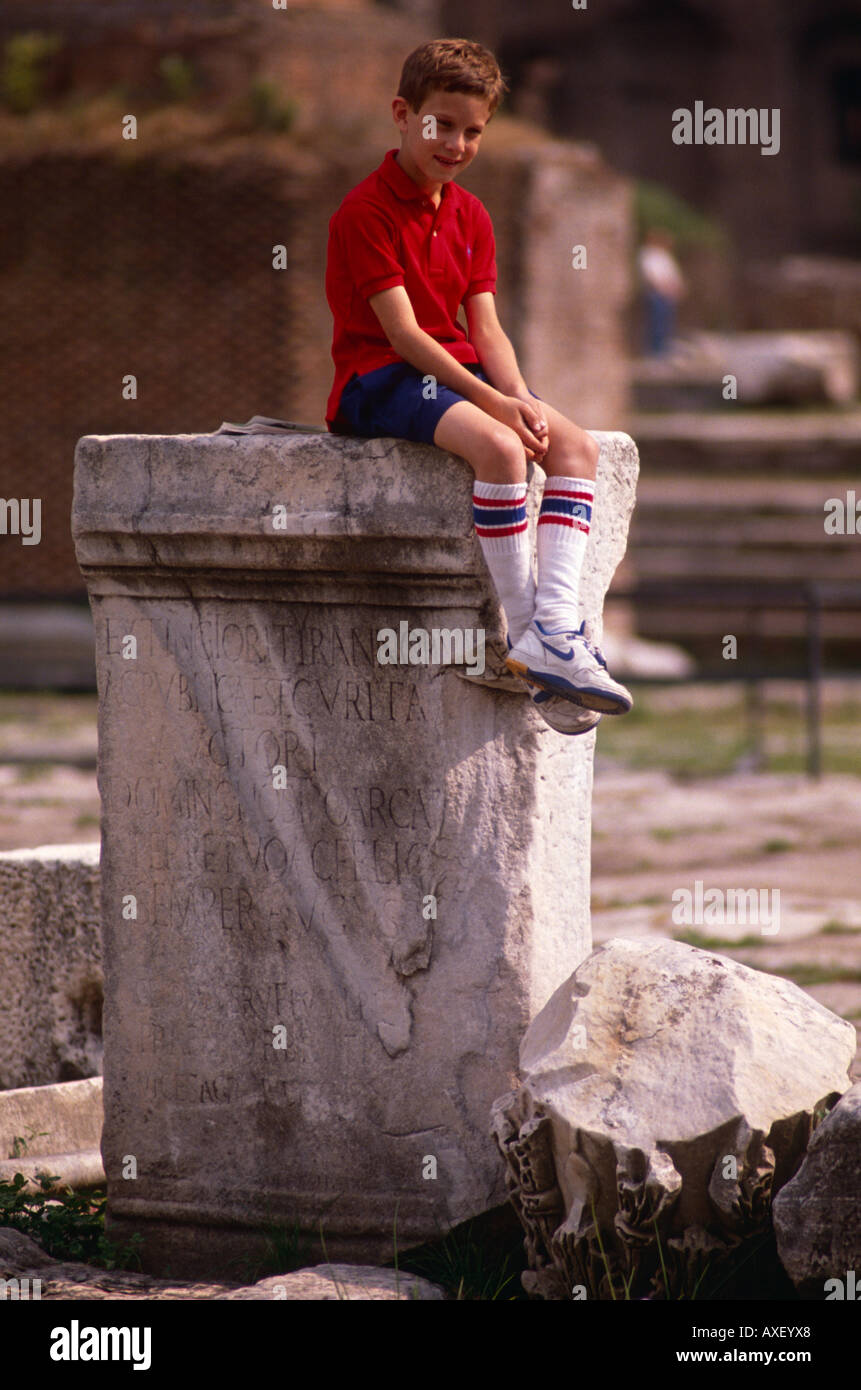 Boy tourist sits on top of an ancient statue pedestal in the ruins of the Roman Forum in central Rome, Italy Stock Photo