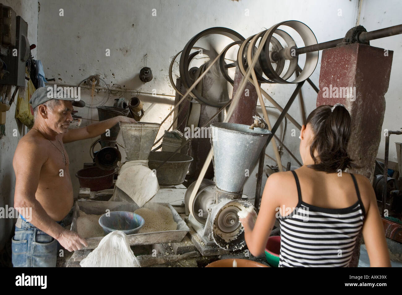 Woman grinding grain into flour at a community molina aka mill in Suchitoto El Salvador while mechanic tends belts and pulleys Stock Photo