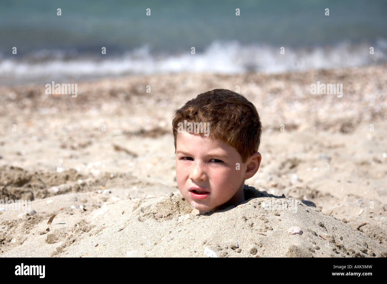 Young boy child head buried up to his neck in sand looking annoyed or