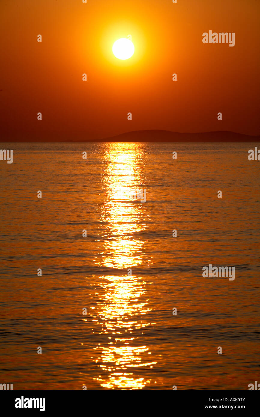 Red orange and gold colours of the sun setting into the sea above island at sunset with reflections in the water in Saronida Att Stock Photo