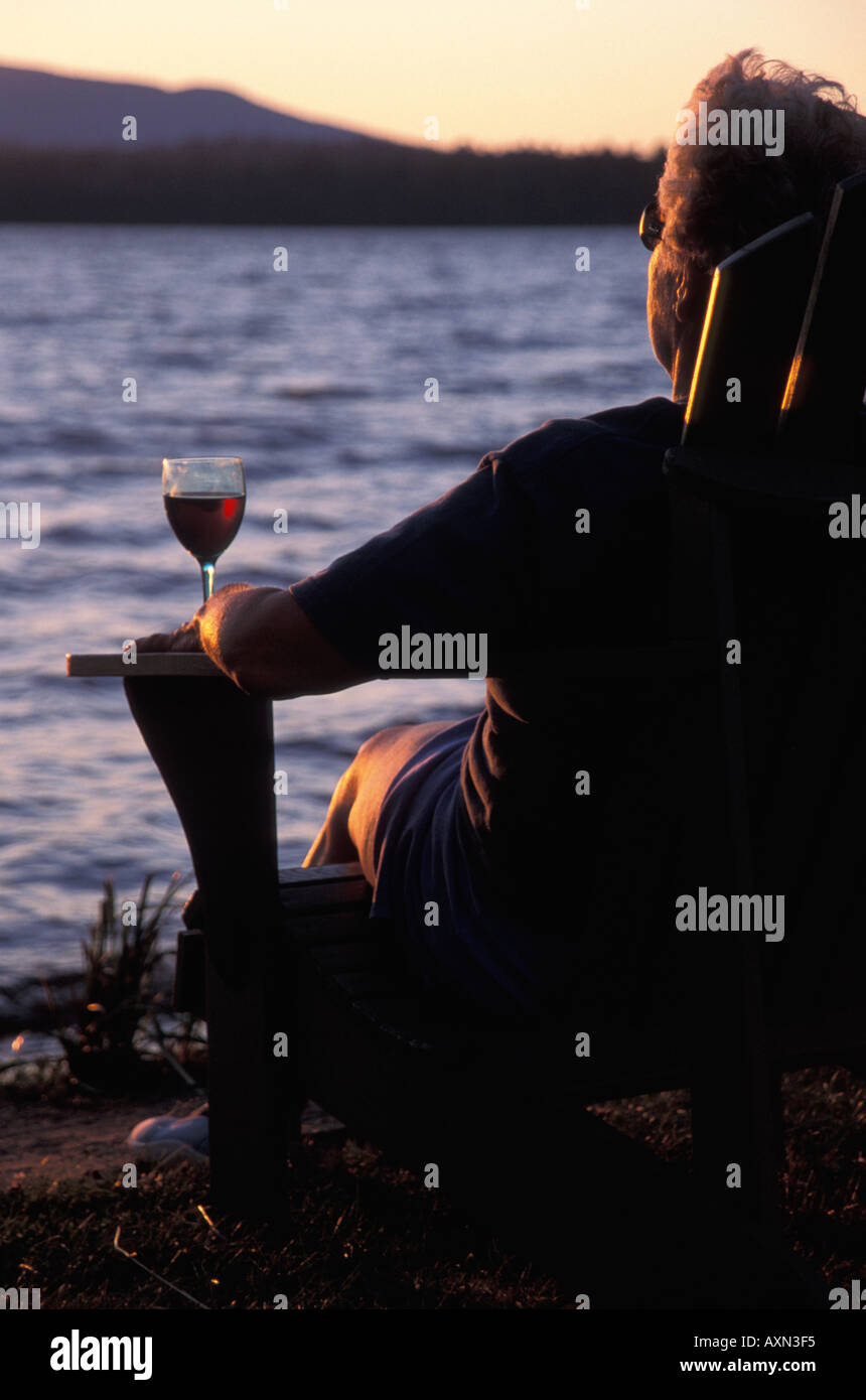 A single older adult woman sits alone near a calm blue lake with a glass of wine contemplating life and watching a sunset. Stock Photo