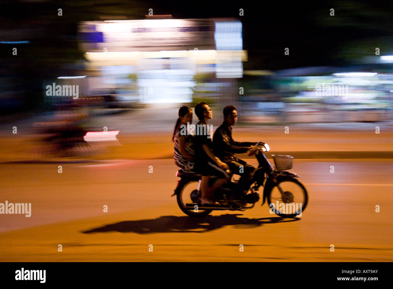 3 people ride one motorcycle past blurred bright lights in Siem Reap, Cambodia near Angkor Wat Stock Photo