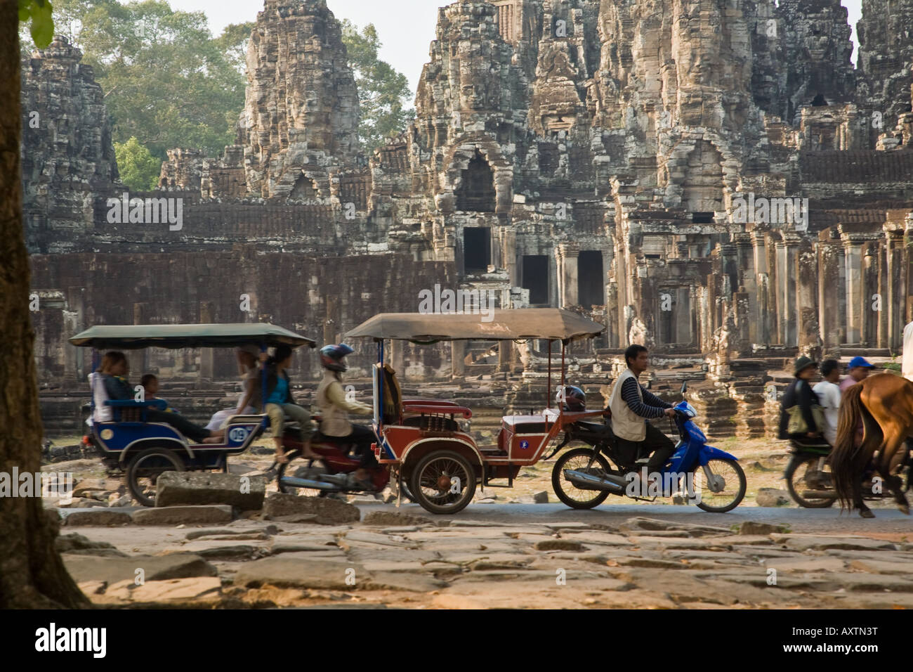 Rickshaws, motorcycles, and horses transport people past the Bayon Temple in  Angkor Thom Stock Photo
