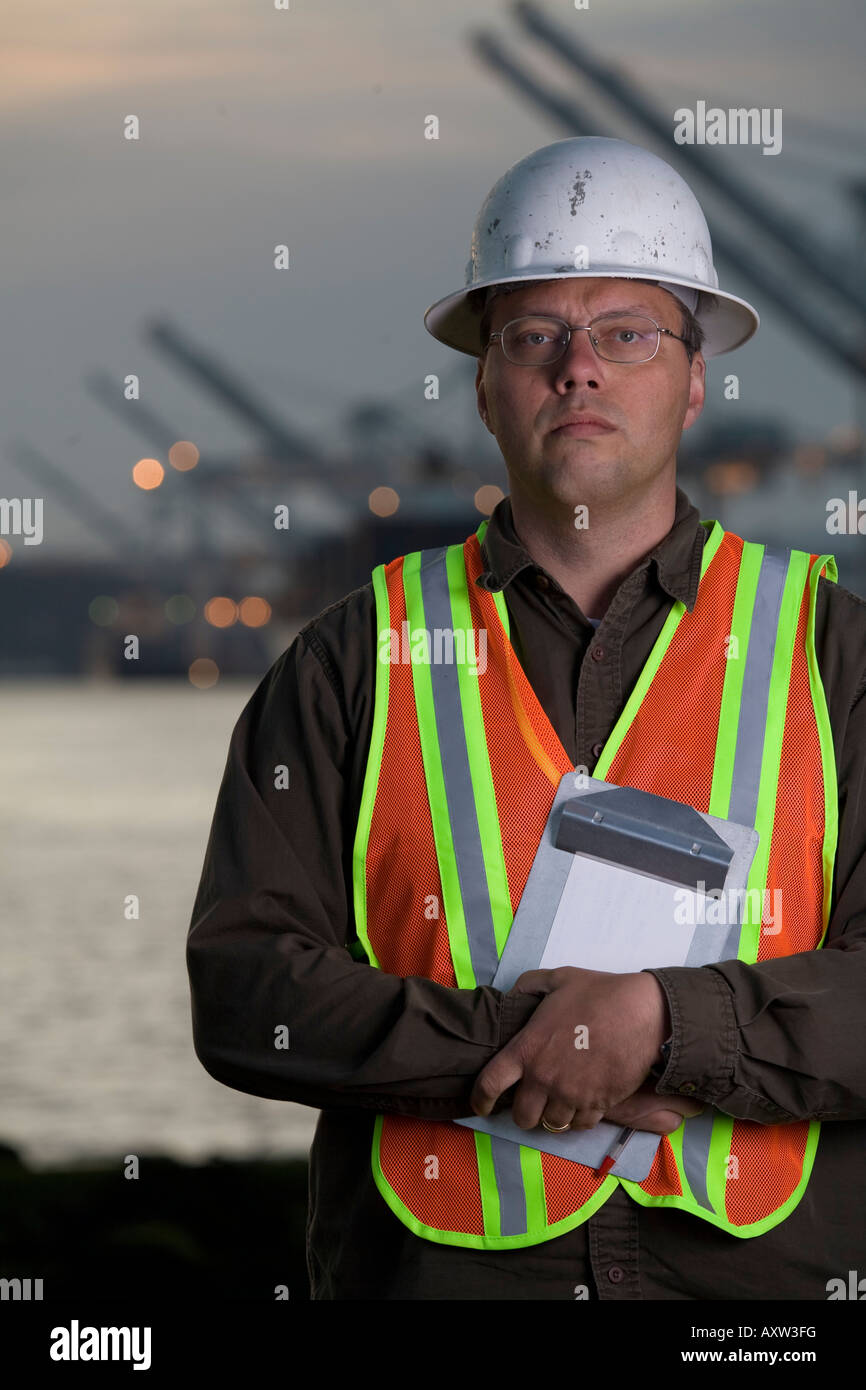 Vertical photo of man with looking to the camera wearing safety vest and hard hat at port with cranes in the back. Stock Photo
