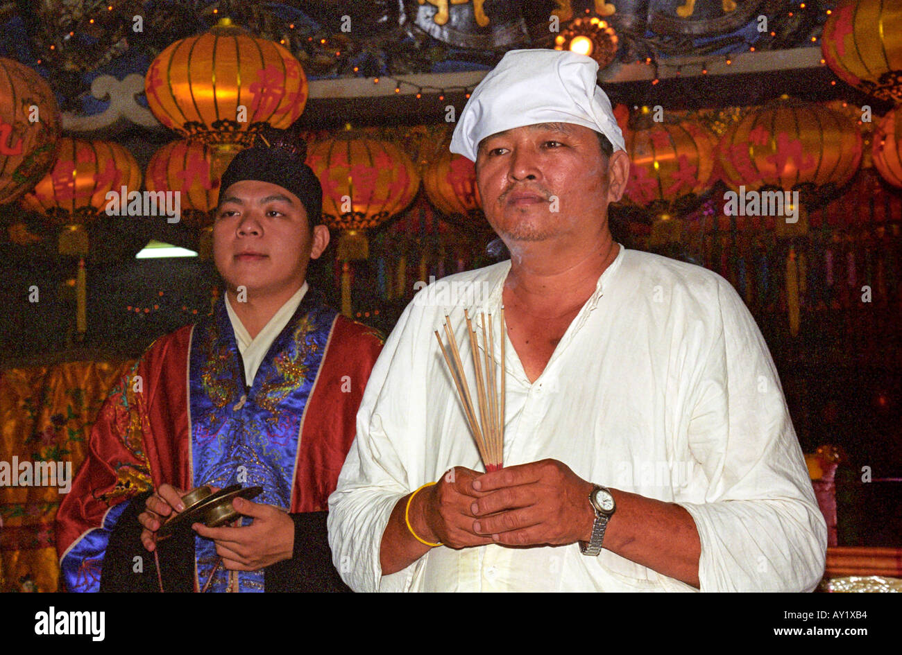 A Toaist devotee dressed in traditional white clothing, holding josticks and praying beside a Taoist priest Stock Photo