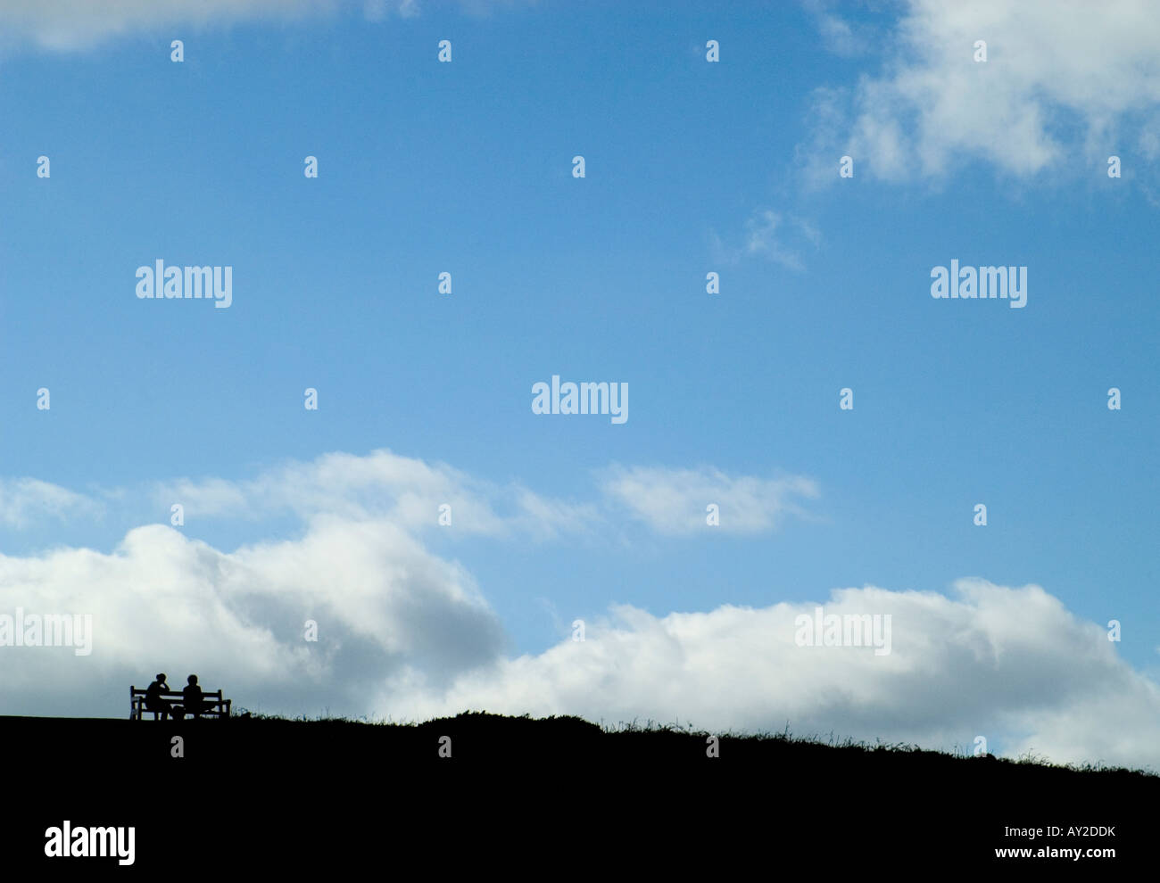 Silhouette of two people sitting on a bench with blue sky and fluffy ...