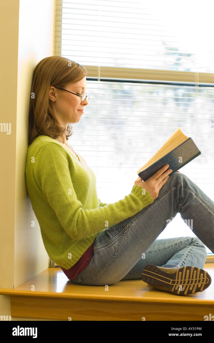 a young woman sitting on a window sill at the library reading a book ...