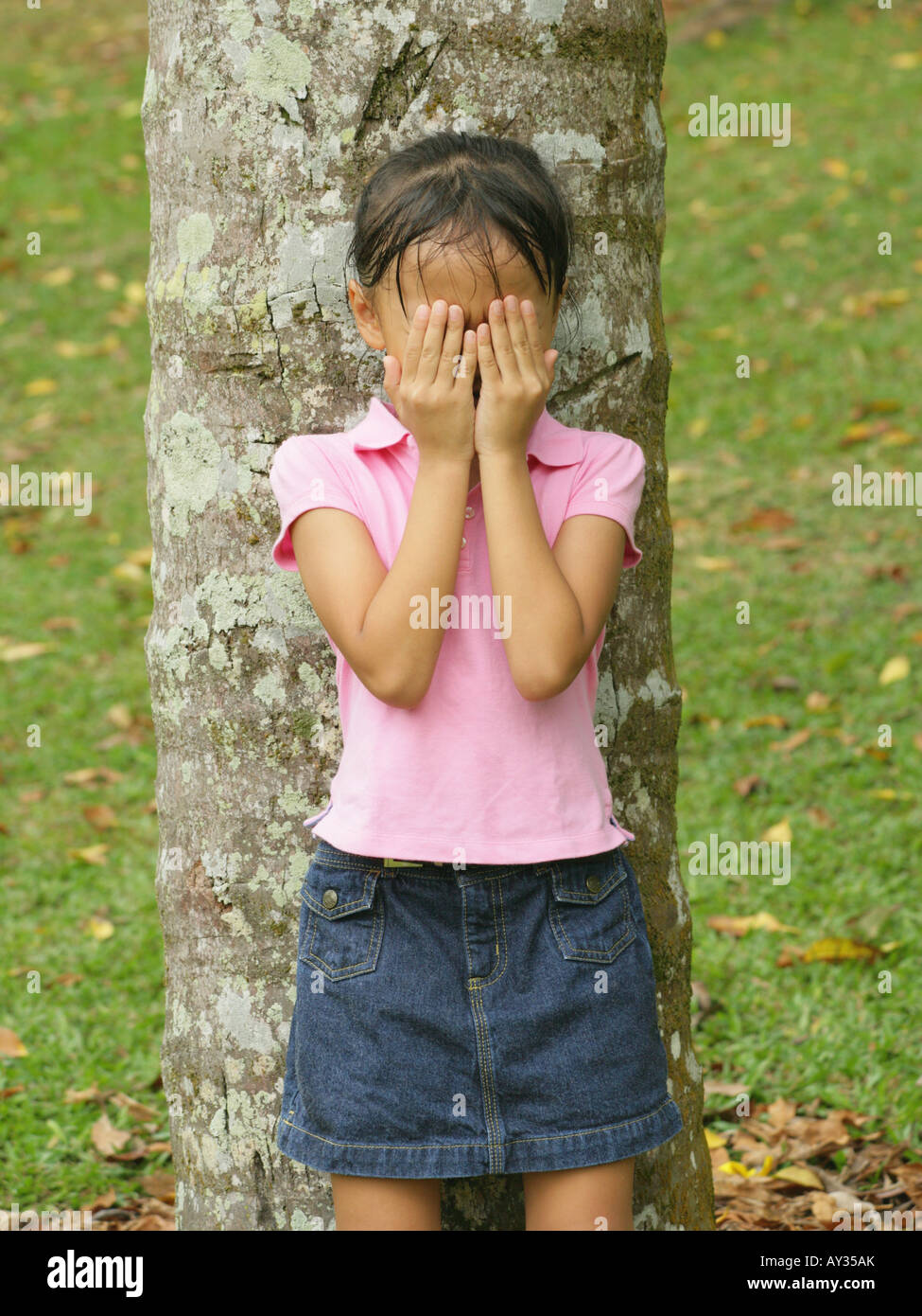 Girl standing in front of a tree trunk and covering her face with her hands Stock Photo