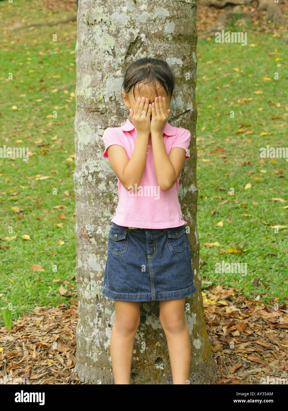 Girl standing in front of a tree trunk and covering her face with her hands Stock Photo