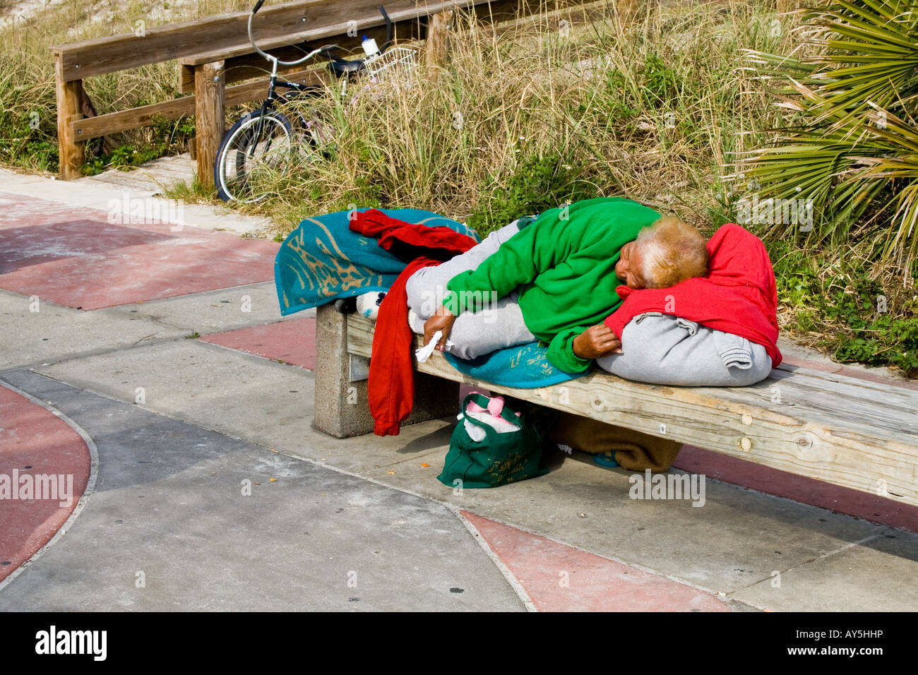 Woman sleeping on a bench in multi-colored clothing next to a sidewalk by a wooden ramp in Jacksonville Beach, Florida, USA Stock Photo