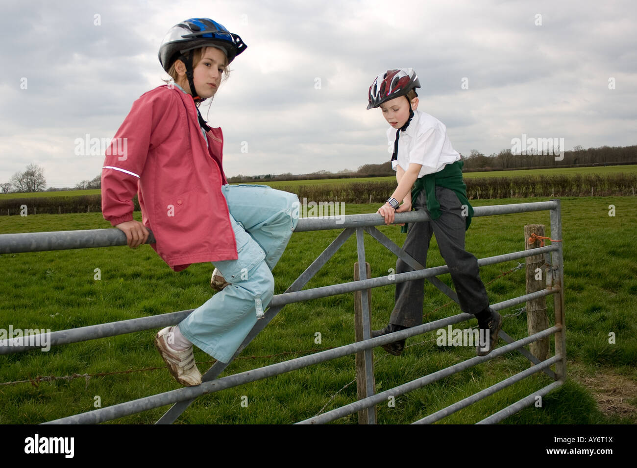 a boy and a girl with cycle helmets playing on a gate in a field Stock Photo