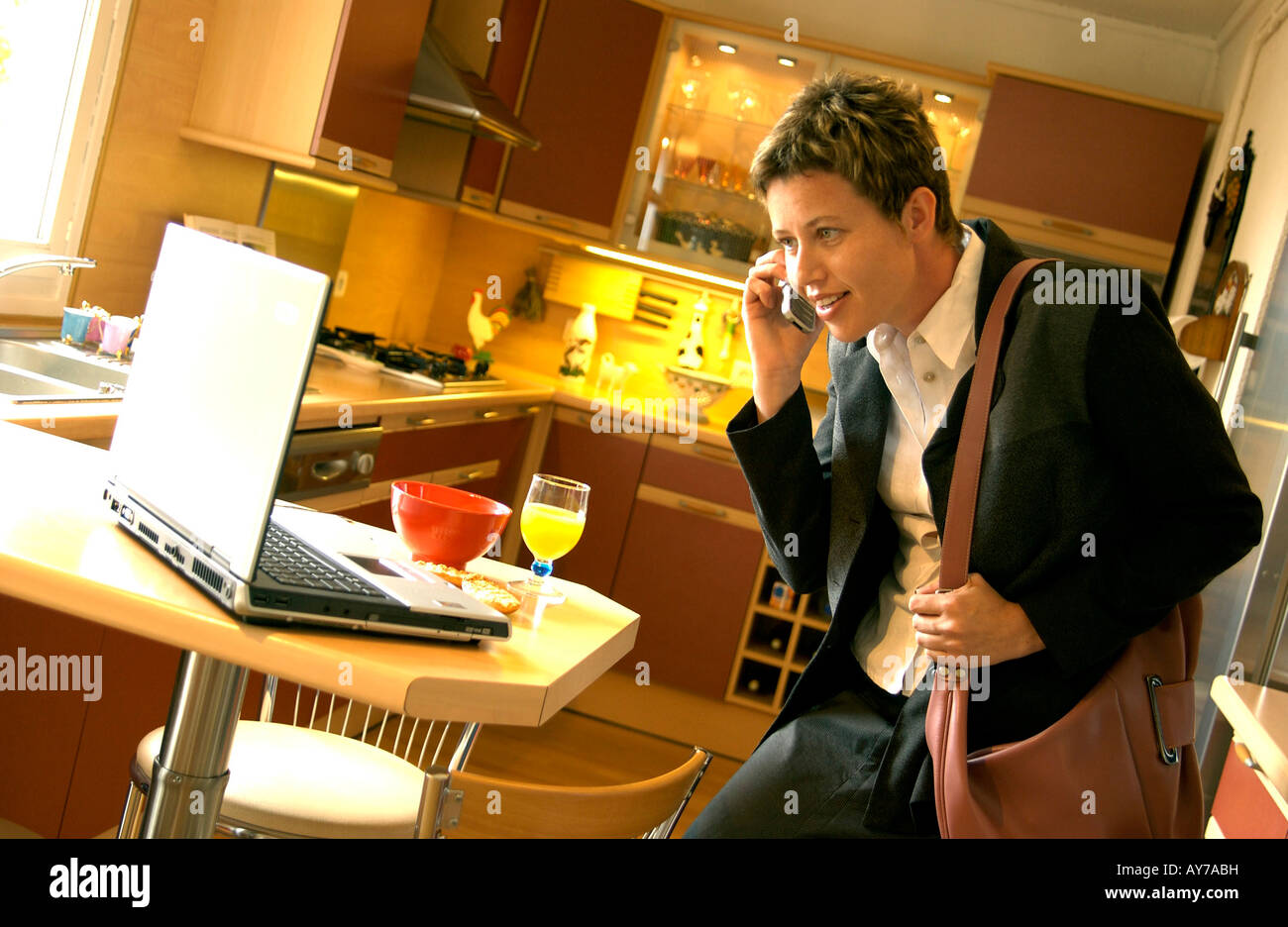 Businesswoman multitasking on phone with computer laptop in the kitchen in the morning rush after breakfast Stock Photo