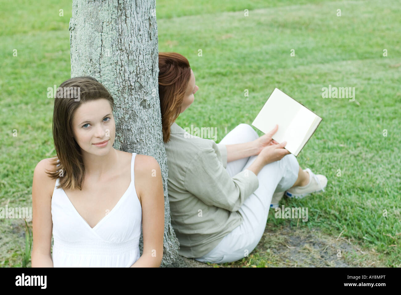 Mother and daughter leaning against tree trunk, woman reading book, teen girl smiling at camera Stock Photo
