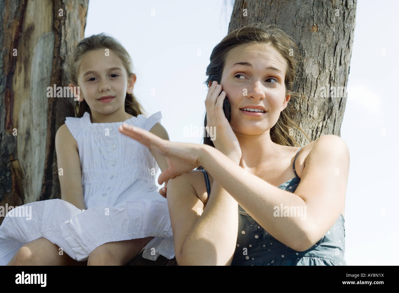 Young woman using cell phone, brushing little sister's hand off her shoulder Stock Photo